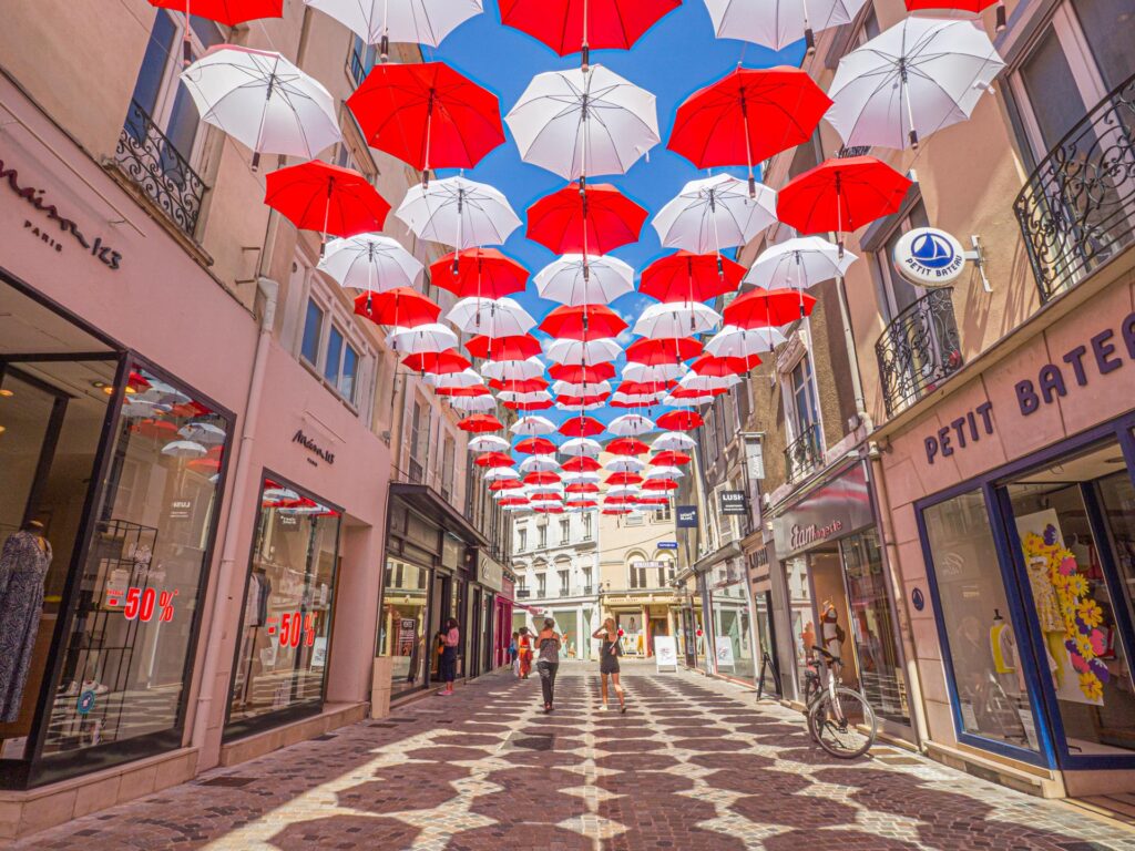 Une photo montrant une installation artistique composée de centaines de parapluies rouge et blanc accrochés à des fils tendus au dessus d’une rue commerçante du Mans. La photo a été prise en 2020 par ParisianGeek, lors de la manifestation “Le Mans sous les parapluies”, qui visait à embellir la ville et à soutenir les commerçants locaux.