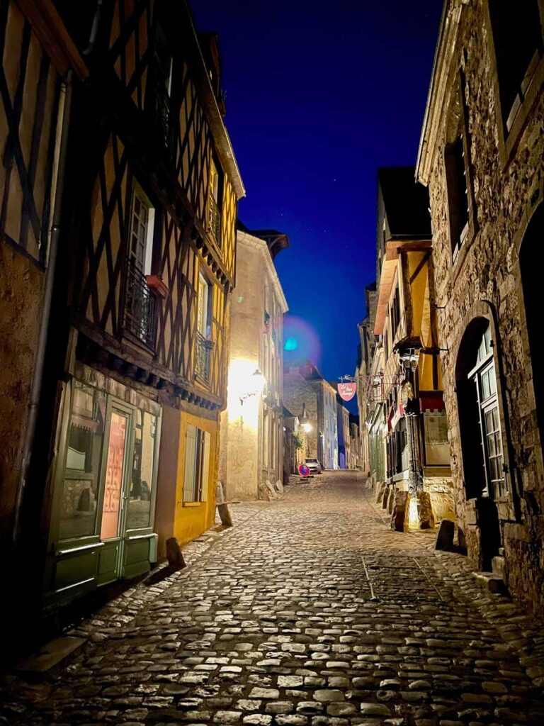 Une photo montrant une rue pavée bordée de maisons à colombages et de lampadaires dans la Cité Plantagenêt du Mans. La rue est déserte et éclairée par la lumière des fenêtres et des réverbères.
