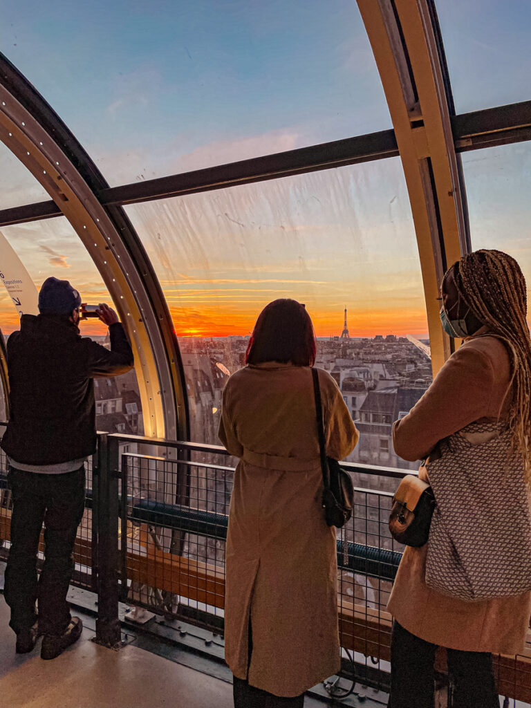 Sur la terrasse du Centre Pompidou, des visiteurs profitent d'une vue panoramique sur Paris. Ils contemplent le coucher du soleil qui colore les toits et les monuments de la ville. Un moment magique et romantique au cœur de la capitale.