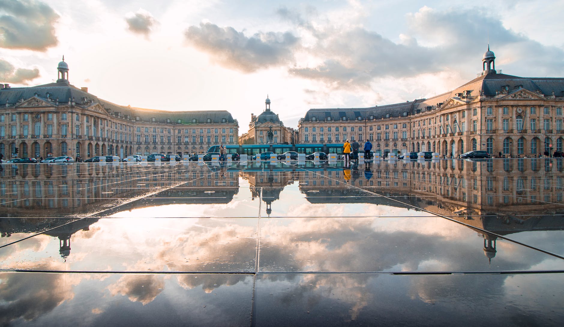 La photo représente le Palais de la Bourse de Bordeaux qui est reflété dans un grand miroir d'eau devant lui. Le bâtiment historique présente une façade de pierre ornée et un tramway passe devant le palais. Le miroir d'eau est parfaitement calme, reflétant le palais de manière claire et nette. Les couleurs dominantes de la photo sont le bleu, le gris et le brun. Il y a également quelques nuages dans le ciel.