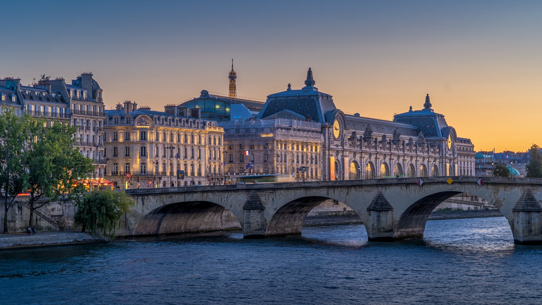 Une photo du pont Royal et du musée d’Orsay illuminés la nuit, vus depuis un quai de Seine à Paris, avec la tour Eiffel illuminée en arrière-plan.