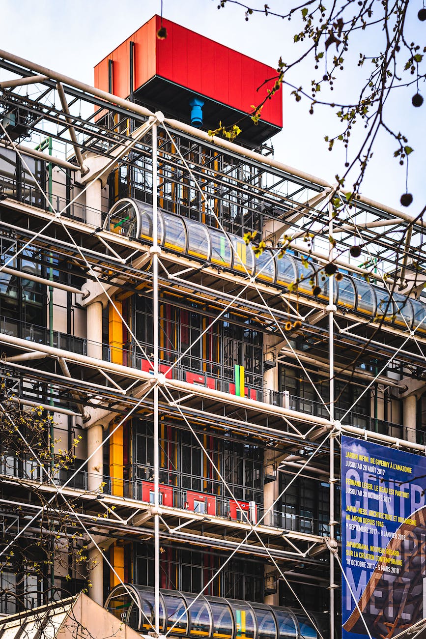 Une photo en gros plan de la façade du Centre Pompidou, qui montre les tuyaux et les escaliers mécaniques de différentes couleurs. On distingue aussi les fenêtres et les panneaux du bâtiment. Un style architectural moderne et original
